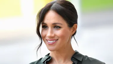 A person with dark hair smiles warmly, wearing a dark green blouse against a light blurred background.
