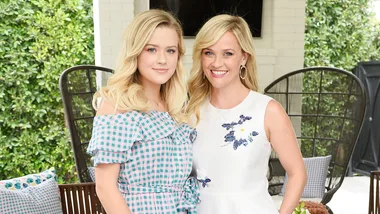 Two women smiling, one in a blue checkered dress and the other in a white floral dress, standing outdoors.