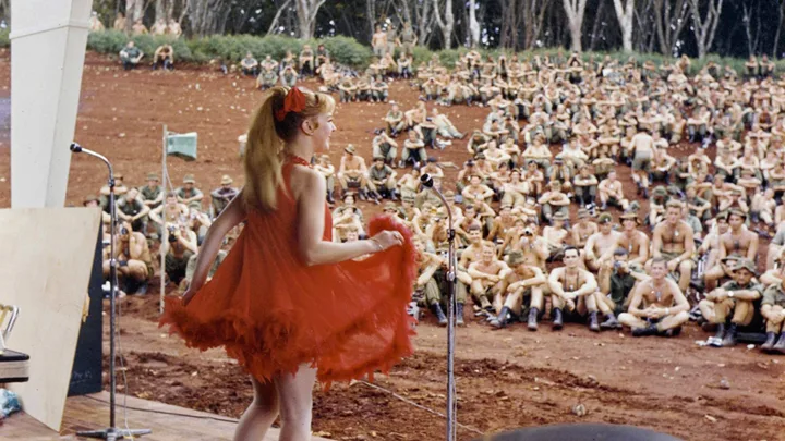 A girl on stage in a red dress performing for soldiers in the war.