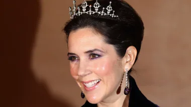 A woman with dark hair smiles, wearing a sparkling tiara and dangling earrings against a neutral background.