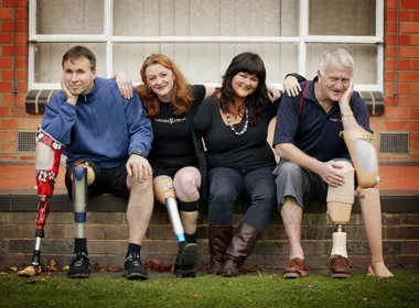 Four people sitting together, three with prosthetic legs, smiling in front of a brick building.