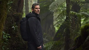 A man in a black coat with a backpack stands in a lush, green forest, glancing back slightly.