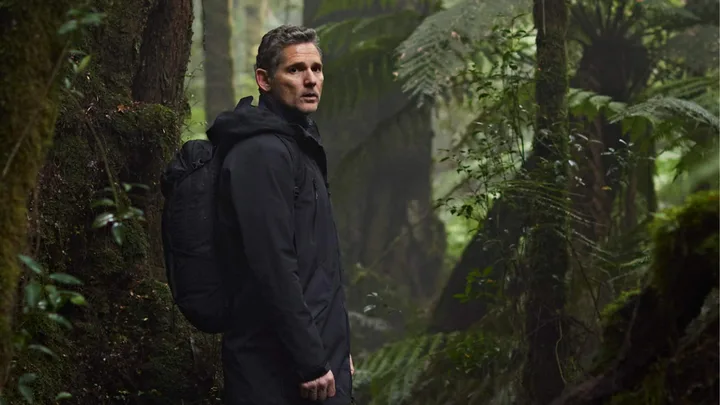 A man in a black coat with a backpack stands in a lush, green forest, glancing back slightly.
