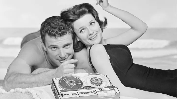 A man and woman on a beach, smiling, with an old reel-to-reel tape recorder in the foreground.