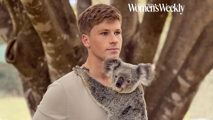 Young man holding a koala against a tree backdrop, featured in The Australian Women's Weekly.