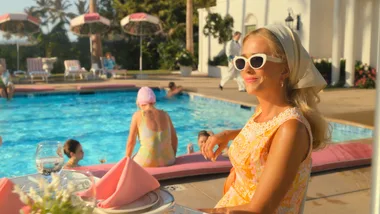 A woman in retro attire with sunglasses and a headscarf sits by a pool, with people and umbrellas in the background.