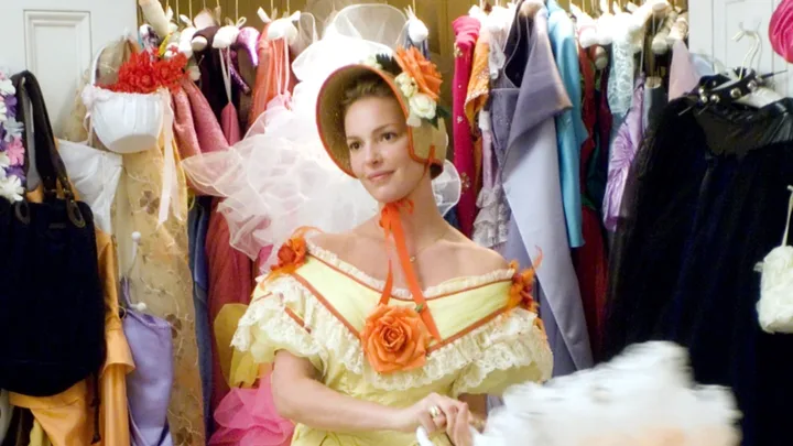 Woman in a yellow and orange dress and hat stands in a closet full of colorful dresses.
