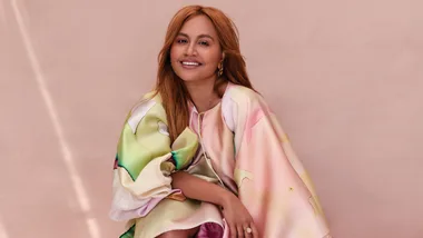 Smiling woman with long hair wearing a colorful dress against a soft pink background.