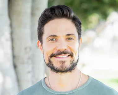 Man with dark hair and beard, wearing a green t-shirt, smiling outdoors with trees in the background.