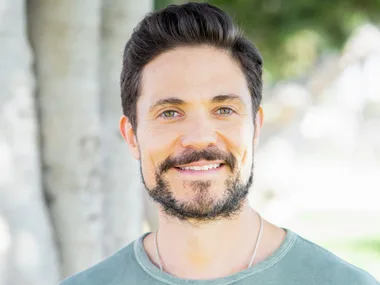 Man with dark hair and beard, wearing a green t-shirt, smiling outdoors with trees in the background.