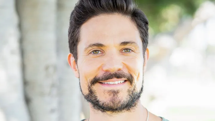 Man with dark hair and beard, wearing a green t-shirt, smiling outdoors with trees in the background.
