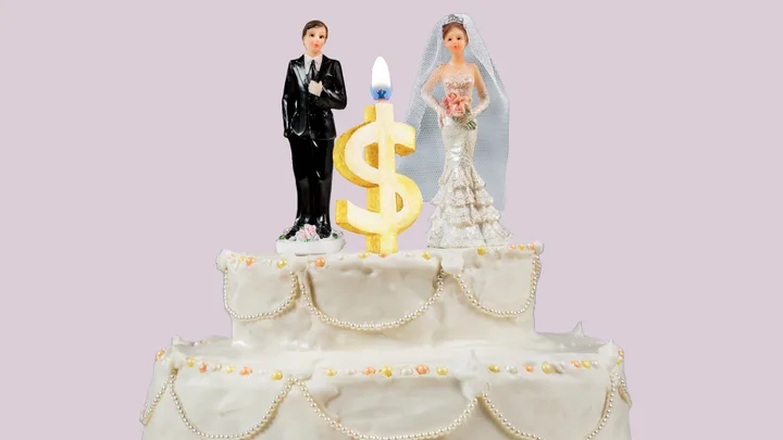 Wedding cake with bride and groom figurines, dollar-sign candle on top, light purple background.