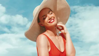 Woman in a red swimsuit and large sun hat smiles under a bright blue sky.