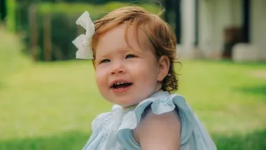 A smiling toddler with red hair and a big bow, wearing a light blue dress, sits on grass with a house in the background.