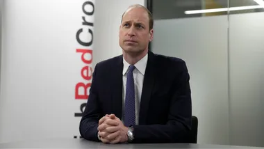 Man in suit with hands clasped, sitting at a table, Red Cross banner in background.