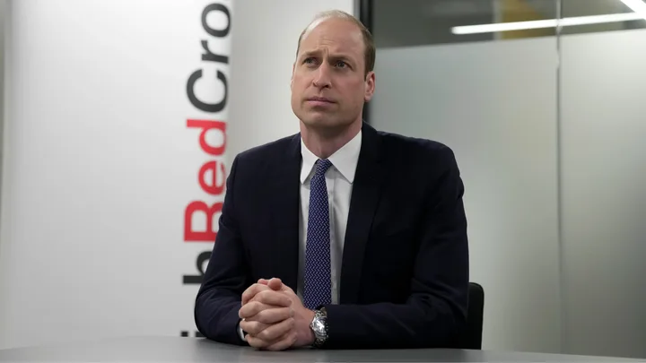 Man in suit with hands clasped, sitting at a table, Red Cross banner in background.