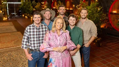 Group of six people posing indoors with rugs and greenery in the background, one woman in a pink dress in front.