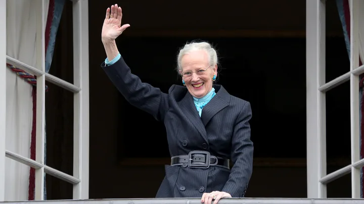 An older woman in a suit waves from a balcony, smiling warmly, with decorative window curtains beside her.