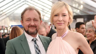 A man and a woman smiling at an event under a tented ceiling, with people in the background.