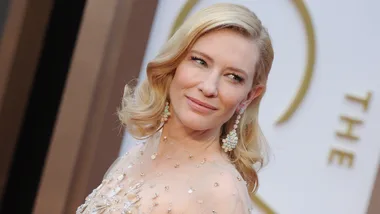 Elegant woman with blonde hair, wearing jeweled earrings and a sparkling dress at an event with a light background.