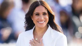 A person smiling, wearing a white blazer and necklace, outdoors in a sunny setting.