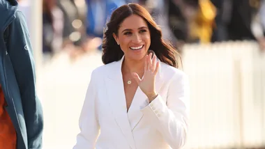 A woman in a white outfit waves and smiles in a bright outdoor setting.
