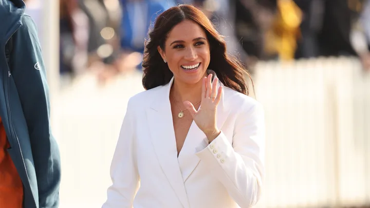 A woman in a white outfit waves and smiles in a bright outdoor setting.