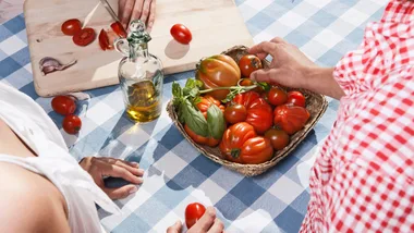 People preparing tomatoes on a checkered tablecloth, with a basket of tomatoes and a bottle of olive oil.