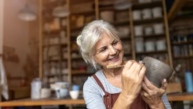 Elderly woman in apron happily painting pottery in workshop with shelves of supplies in background.