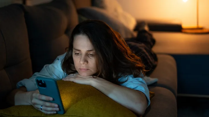 Woman lying on sofa, looking at smartphone screen, with a lamp in the background.