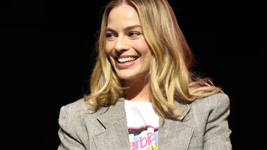 A woman with long blonde hair smiles, wearing a gray blazer over a white shirt, against a dark background.