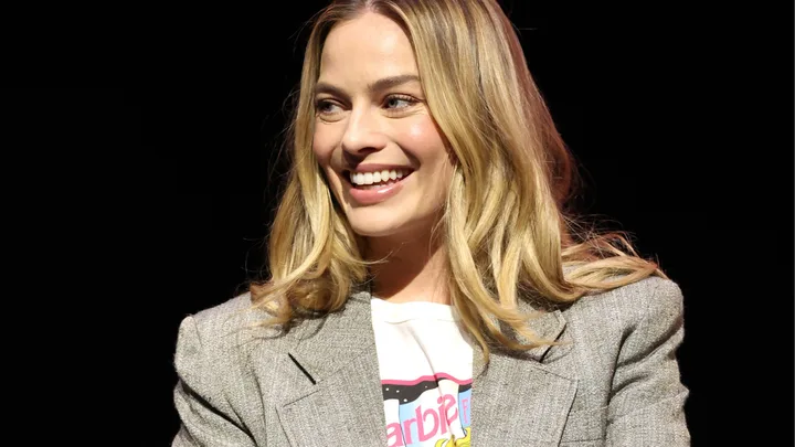 A woman with long blonde hair smiles, wearing a gray blazer over a white shirt, against a dark background.