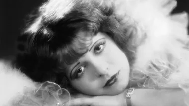 Portrait of a woman with curly hair and a dreamy expression, resting her head on folded arms, wearing a fluffy garment.