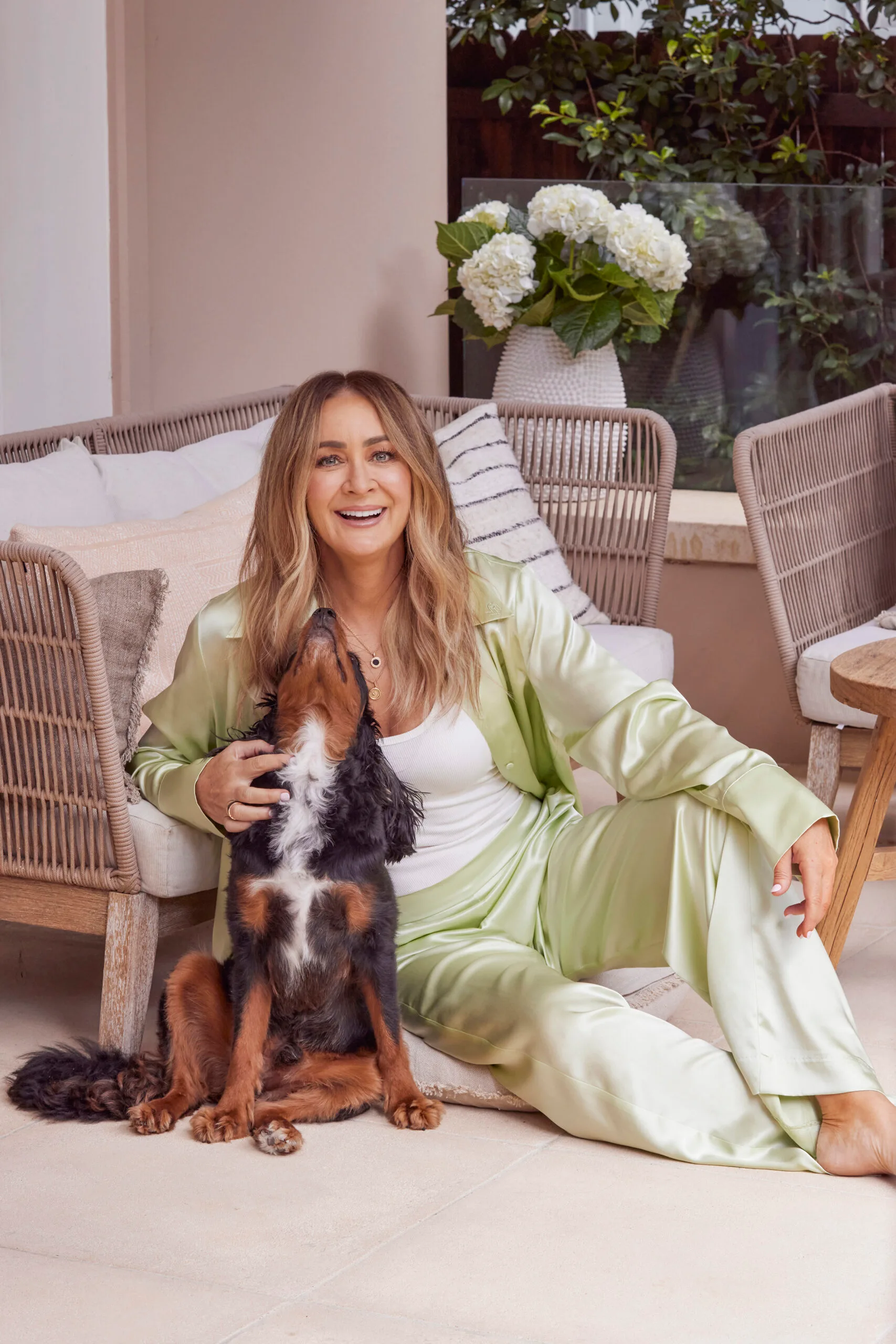 Woman in green silk outfit sitting with a dog indoors, smiling, near a wicker chair and white flowers.