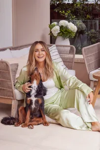 Woman in green silk outfit sitting with a dog indoors, smiling, near a wicker chair and white flowers.