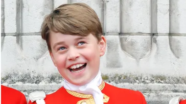 A young child in a red and gold uniform smiles broadly in front of a stone wall.