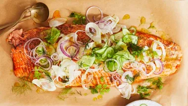 Roasted salmon topped with sliced fennel, red onion, cucumber, dill, and herbs on parchment paper.