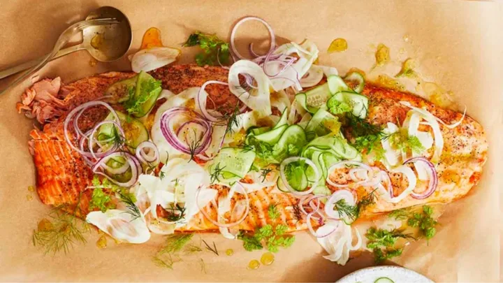 Roasted salmon topped with sliced fennel, red onion, cucumber, dill, and herbs on parchment paper.