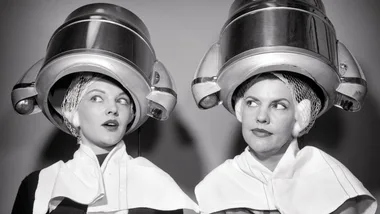 Two women sitting under vintage hairdryers, wearing hairnets and capes, exchange amused looks in a salon.