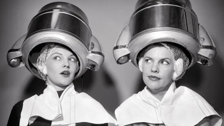 Two women sitting under vintage hairdryers, wearing hairnets and capes, exchange amused looks in a salon.
