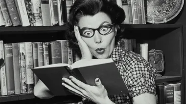 A woman with exaggerated glasses reads a book, appearing surprised, in front of a bookshelf filled with books.