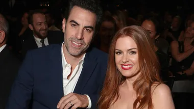 A man in a blue suit and a woman with red hair in a gown, smiling at an event with people in the background.