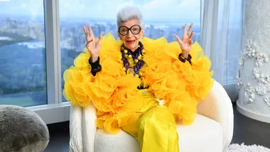 Elderly woman in vibrant yellow ruffled outfit and large glasses, smiling with hands raised, seated in a bright room.