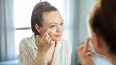 A woman applying face cream in front of a mirror, smiling gently.