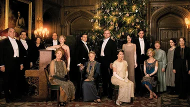 Cast of "Downton Abbey" in formal attire gathered around a decorated Christmas tree inside a grand room.