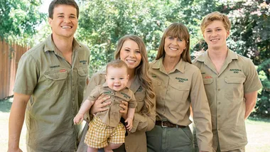 A smiling family of five in khaki Australia Zoo uniforms stands outdoors, holding a baby.