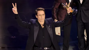 Man in a suit making a peace sign on stage, holding award, celebrating.