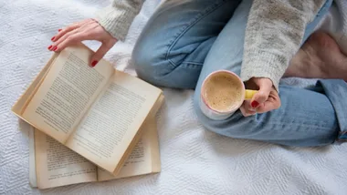 Person sitting on bed in jeans, holding coffee, reading a book with pages slightly open.