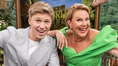 Two smiling people in a jungle-themed setting, one in a light suit and the other in a green dress, in front of a show sign.