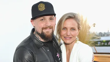 A man in a black leather jacket with a beard and a woman in a white outfit smiling, outdoors with plants in the background.